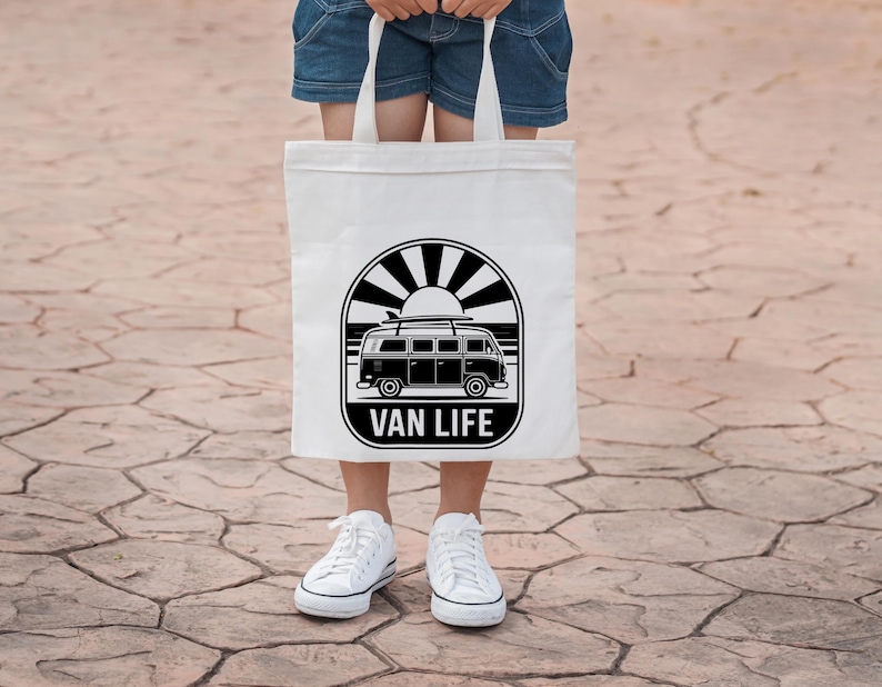 May include: A white tote bag featuring a black and white graphic of a vintage van with surfboards, the sun, and the words "VAN LIFE". The bag is held by a person wearing denim shorts and white sneakers.