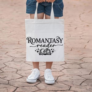 May include: A white tote bag with the words "ROMANTASY reader" in black script. Below the text is a graphic of an open book with a rose and stars. The bag is held by a person wearing denim shorts and white sneakers.