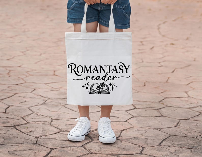 May include: A white tote bag with the words "ROMANTASY reader" in black script. Below the text is a graphic of an open book with a rose and stars. The bag is held by a person wearing denim shorts and white sneakers.