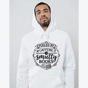 May include: White hoodie with a drawstring and a front pocket. The black graphic reads "Fueled by Caffeine and Smutty Books" with coffee cup, coffee pot, and book illustrations. The hoodie is worn by a person.