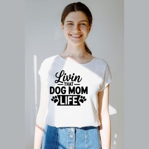 May include: White t-shirt with the text "Livin' that Dog Mom Life" in black script and paw print graphics. The shirt is a crew neck style and is being worn.