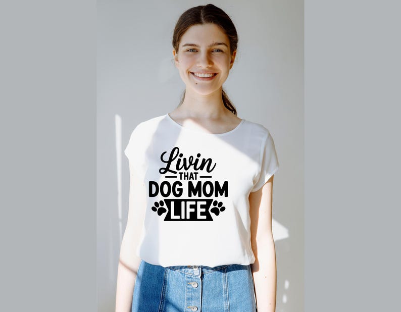 May include: White t-shirt with the text "Livin' that Dog Mom Life" in black script and paw print graphics. The shirt is a crew neck style and is being worn.