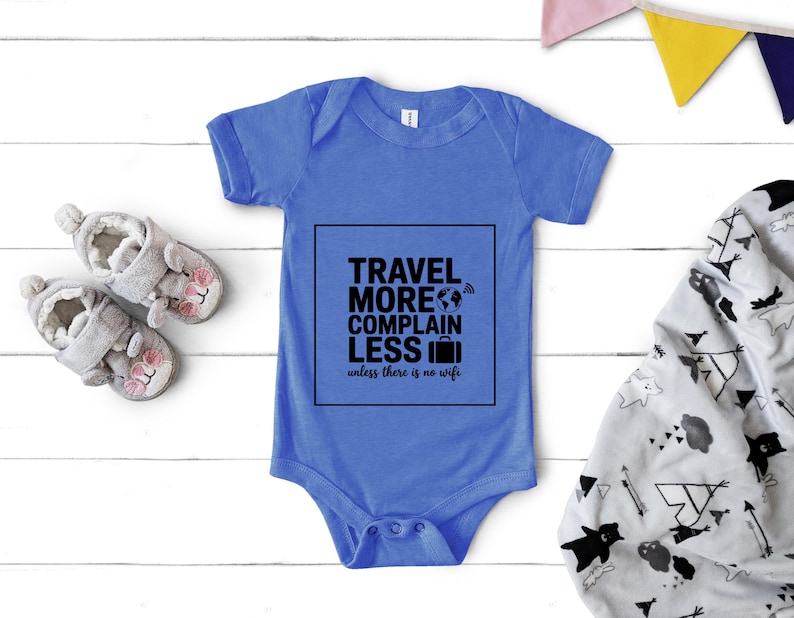 May include: A blue baby onesie with the text "TRAVEL MORE COMPLAIN LESS unless there is no wifi" in a black square. The onesie is next to a pair of gray baby shoes and a patterned blanket.