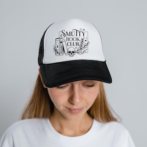 May include: A black and white trucker hat with the text "Smutty Book Club" in a decorative frame. The design includes illustrations of books, a skull, and foliage. The hat has a black brim and mesh back.