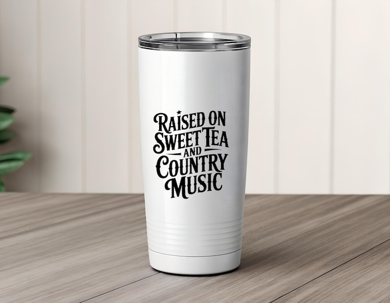 May include: A white insulated tumbler with a silver rim. The tumbler features the black text "Raised on Sweet Tea and Country Music" in a decorative font. The tumbler is designed to keep beverages hot or cold.