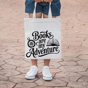 May include: A white tote bag with the text "Books are my Adventure" in black script, featuring a compass and an open book graphic. The bag is held by a person wearing denim shorts and white sneakers.