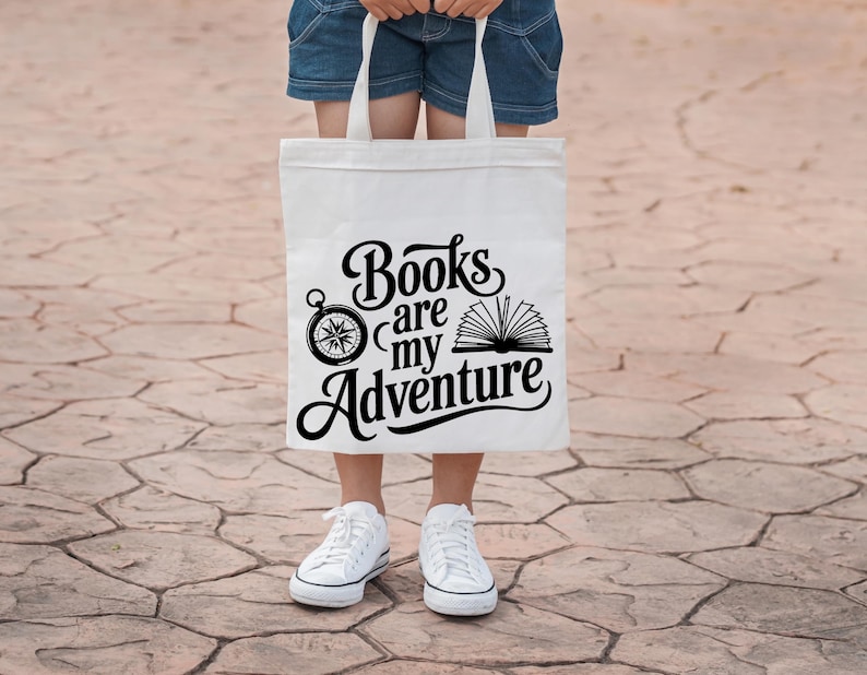 May include: A white tote bag with the text "Books are my Adventure" in black script, featuring a compass and an open book graphic. The bag is held by a person wearing denim shorts and white sneakers.