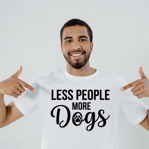 May include: White t-shirt with the text "LESS PEOPLE MORE Dogs" in black. The word "Dogs" features a paw print graphic. The person in the image is pointing at the shirt.