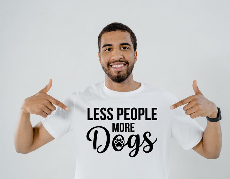 May include: White t-shirt with the text "LESS PEOPLE MORE Dogs" in black. The word "Dogs" features a paw print graphic. The person in the image is pointing at the shirt.