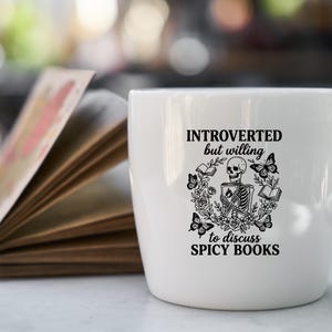 May include: White ceramic mug with the text "INTROVERTED but willing to discuss SPICY BOOKS" and a graphic of a skeleton, books, and butterflies. An open book is in the background.