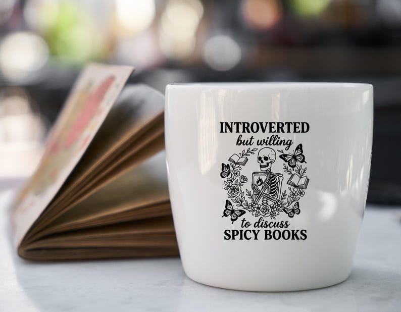 May include: White ceramic mug with the text "INTROVERTED but willing to discuss SPICY BOOKS" and a graphic of a skeleton, books, and butterflies. An open book is in the background.