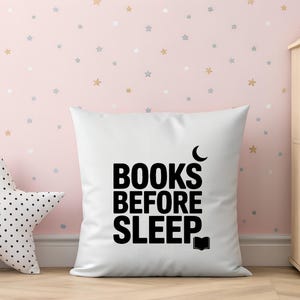 May include: White square pillow with the text "BOOKS BEFORE SLEEP" in black. A crescent moon and an open book graphic are included. A star-shaped pillow with black polka dots is on the left. The background is a pink wall with star decals.