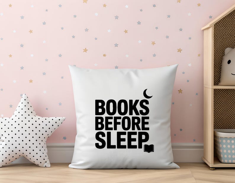 May include: White square pillow with the text "BOOKS BEFORE SLEEP" in black. A crescent moon and an open book graphic are included. A star-shaped pillow with black polka dots is on the left. The background is a pink wall with star decals.