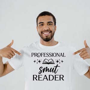 May include: White t-shirt with the text "PROFESSIONAL smut READER" in black. The text is arranged with the word "smut" in a larger, stylized font. The design includes a book graphic and star accents. The person in the image is pointing at the shirt.