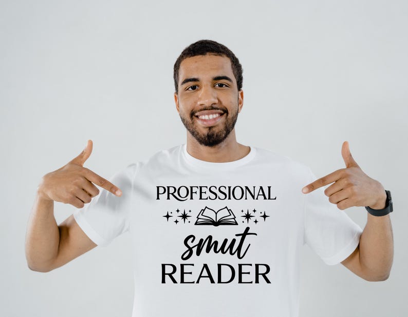 May include: White t-shirt with the text "PROFESSIONAL smut READER" in black. The text is arranged with the word "smut" in a larger, stylized font. The design includes a book graphic and star accents. The person in the image is pointing at the shirt.
