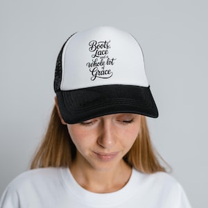 May include: A black and white trucker hat with the text "Boots, Lace and a whole lot of Grace" in a script font. The hat has a white front panel and a black brim and mesh back.