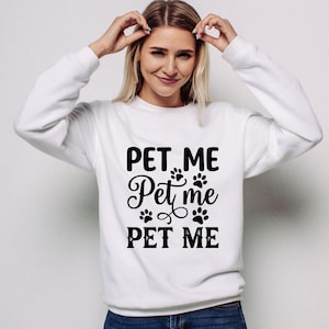 May include: White sweatshirt with the text "PET ME" in a bold, black font, with paw prints and decorative swirls. The sweatshirt is a casual style, suitable for pet lovers.