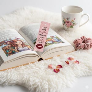 May include: An open book with a pink bookmark that reads "I still read fairytales they are just Duter now" and a lip graphic. A white mug with pink roses and a pink scrunchie are also present. The book is on a white fluffy surface.
