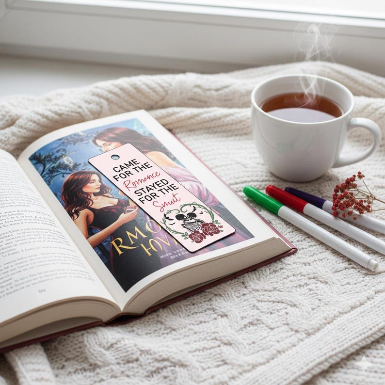 May include: An open book with a bookmark that reads "Came for the Romance, Stayed for the Smut." The bookmark features a skull, roses, and a heart design. A white mug of tea and pens are nearby.