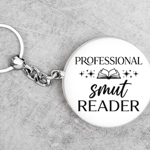 May include: A white keychain with the text "PROFESSIONAL smut READER" in black. The keychain has a silver ring and chain. The design includes an open book and stars.