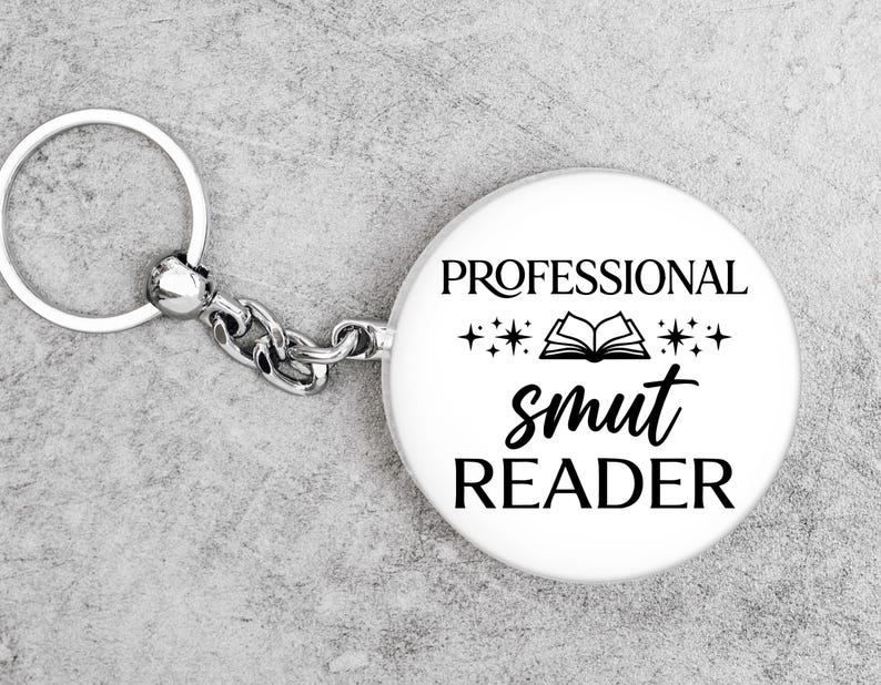 May include: A white keychain with the text "PROFESSIONAL smut READER" in black. The keychain has a silver ring and chain. The design includes an open book and stars.