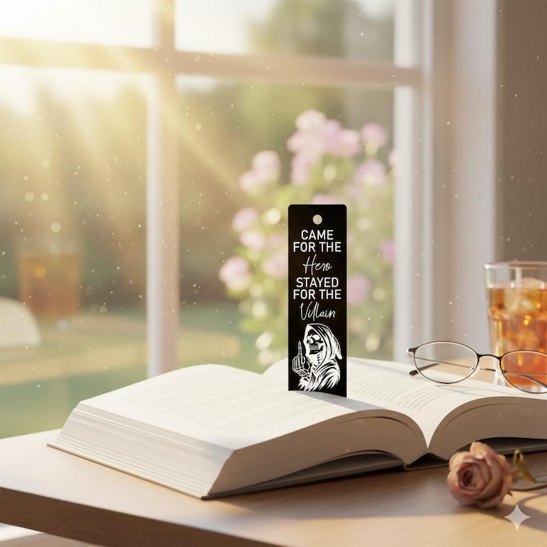 May include: A black bookmark with the text "Came for the Hero, Stayed for the Villain" and a Grim Reaper illustration. The bookmark is inserted into an open book on a wooden table, with a glass of iced tea and glasses in the background.