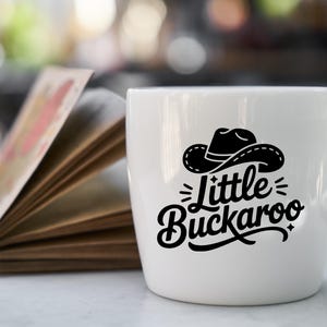 May include: A white ceramic mug with a black graphic of a cowboy hat and the words "Little Buckaroo". The mug is on a white surface next to an open book with colorful pages. The background is blurred.