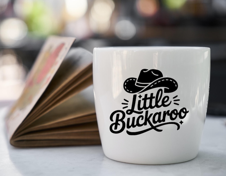 May include: A white ceramic mug with a black graphic of a cowboy hat and the words "Little Buckaroo". The mug is on a white surface next to an open book with colorful pages. The background is blurred.