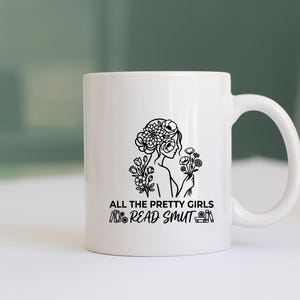 May include: White ceramic mug with a black line drawing of a woman with flowers in her hair and holding flowers. The text "ALL THE PRETTY GIRLS READ SMUT" is printed below the illustration. The mug has a curved handle.