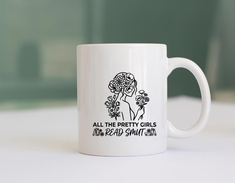 May include: White ceramic mug with a black line drawing of a woman with flowers in her hair and holding flowers. The text "ALL THE PRETTY GIRLS READ SMUT" is printed below the illustration. The mug has a curved handle.