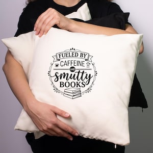 May include: Cream-colored pillow with the text "Fueled by Caffeine and Smutty Books" in a black, decorative design. The design includes coffee cup, coffee pot, and book illustrations. The pillow is held by a person wearing a black shirt.