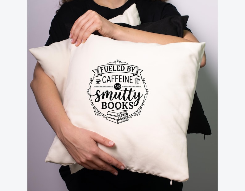 May include: Cream-colored pillow with the text "Fueled by Caffeine and Smutty Books" in a black, decorative design. The design includes coffee cup, coffee pot, and book illustrations. The pillow is held by a person wearing a black shirt.