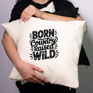 May include: Cream-colored pillow with the text "BORN Country RAISED WILD" in black, stylized lettering. The pillow is held by a person wearing a black shirt. The pillow is square and appears to be made of a soft fabric.