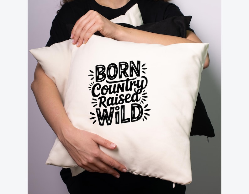 May include: Cream-colored pillow with the text "BORN Country RAISED WILD" in black, stylized lettering. The pillow is held by a person wearing a black shirt. The pillow is square and appears to be made of a soft fabric.