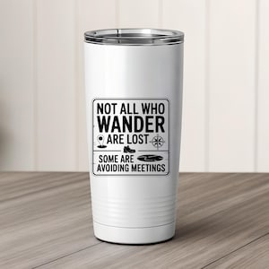 May include: White travel tumbler with a silver rim. The tumbler features a black graphic with the text "NOT ALL WHO WANDER ARE LOST. SOME ARE AVOIDING MEETINGS" and icons of a map pin, boot, compass, and lake. The tumbler is on a wooden surface.