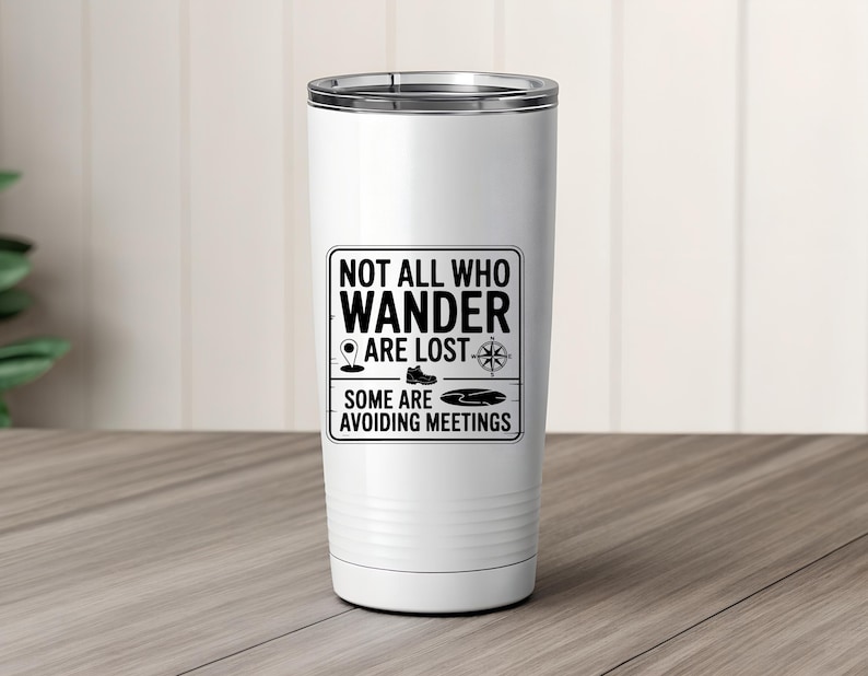 May include: White travel tumbler with a silver rim. The tumbler features a black graphic with the text "NOT ALL WHO WANDER ARE LOST. SOME ARE AVOIDING MEETINGS" and icons of a map pin, boot, compass, and lake. The tumbler is on a wooden surface.