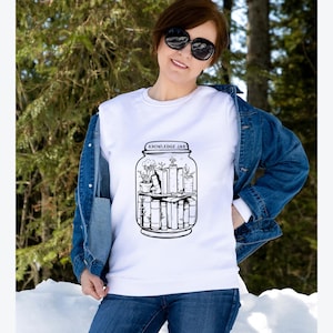 May include: White sweatshirt with a black and white graphic of a jar labeled "KNOWLEDGE JAR" containing books and plants. The sweatshirt is worn with a denim jacket and blue jeans.