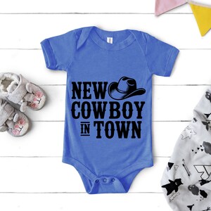 May include: A blue baby onesie with the text "NEW COWBOY IN TOWN" and a cowboy hat graphic. The onesie is displayed with a pair of gray baby shoes and a patterned blanket.