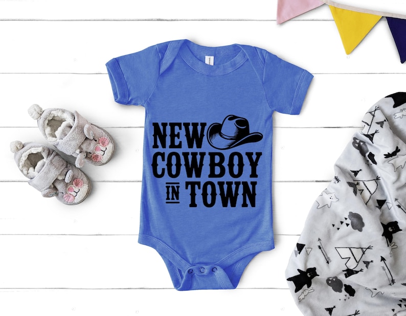 May include: A blue baby onesie with the text "NEW COWBOY IN TOWN" and a cowboy hat graphic. The onesie is displayed with a pair of gray baby shoes and a patterned blanket.