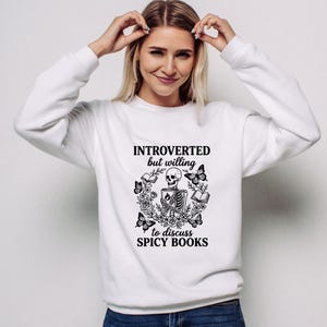 May include: White sweatshirt with the text "INTROVERTED but willing to discuss SPICY BOOKS" and a black graphic of a skeleton, butterflies, flowers, and books. The sweatshirt is a crew neck style.