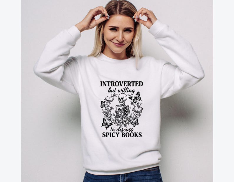May include: White sweatshirt with the text "INTROVERTED but willing to discuss SPICY BOOKS" and a black graphic of a skeleton, butterflies, flowers, and books. The sweatshirt is a crew neck style.