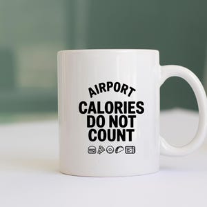 May include: A white ceramic mug with the text "AIRPORT CALORIES DO NOT COUNT" in black. The mug features small food icons below the text, including a burger, pizza slice, donut, taco, and a boarding pass.