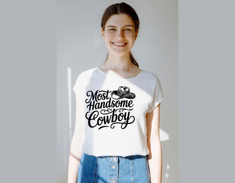 May include: White t-shirt with the text "Most Handsome Cowboy" in a black, stylized font, and a cowboy hat graphic. The shirt is paired with blue denim shorts.