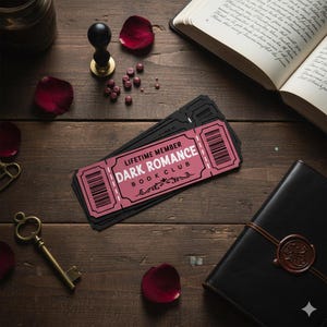 May include: A stack of black tickets with a pink rectangle that reads "LIFETIME MEMBER DARK ROMANCE BOOK CLUB" on a wooden surface. Other items include a book, a key, rose petals, and a journal.
