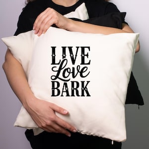 May include: Cream-colored square pillow with the words "LIVE Love BARK" in black script. The pillow is held by a person wearing a black shirt. A black pillow is also visible.