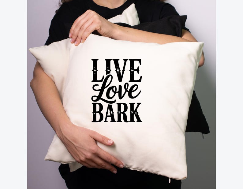 May include: Cream-colored square pillow with the words "LIVE Love BARK" in black script. The pillow is held by a person wearing a black shirt. A black pillow is also visible.