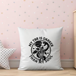 May include: White square pillow with a black graphic design. The design features a skeleton hand holding a rose, a snake, and the text "MY TBR IS DARKER THAN MY SOUL" in a circular arrangement. The pillow is on a wooden surface.