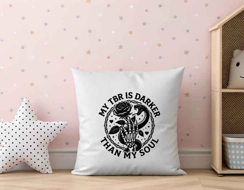 May include: White square pillow with a black graphic design. The design features a skeleton hand holding a rose, a snake, and the text "MY TBR IS DARKER THAN MY SOUL" in a circular arrangement. The pillow is on a wooden surface.
