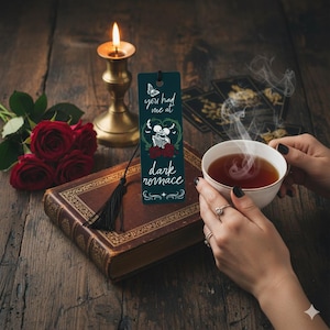 May include: A dark teal bookmark with the text "you had me at dark romance" and a skeleton couple design. The bookmark is on a vintage book with a black tassel, next to red roses, a lit candle, and a cup of tea.