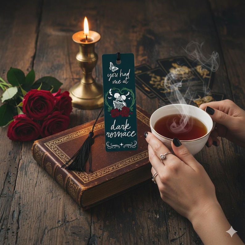 May include: A dark teal bookmark with the text "you had me at dark romance" and a skeleton couple design. The bookmark is on a vintage book with a black tassel, next to red roses, a lit candle, and a cup of tea.